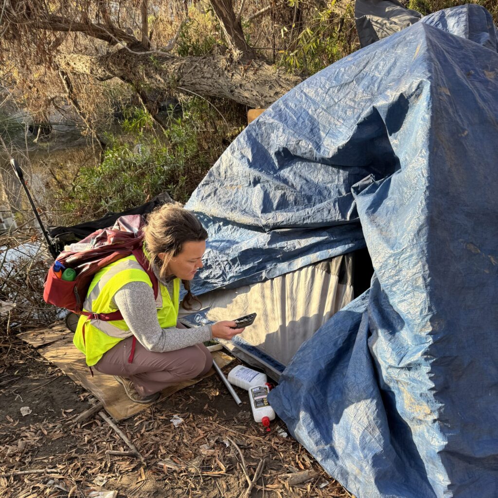 Census Interviewing Person Experiencing Homelessness A volunteer crouches by a tent in the riverbed to interview a person experiencing homelessness.