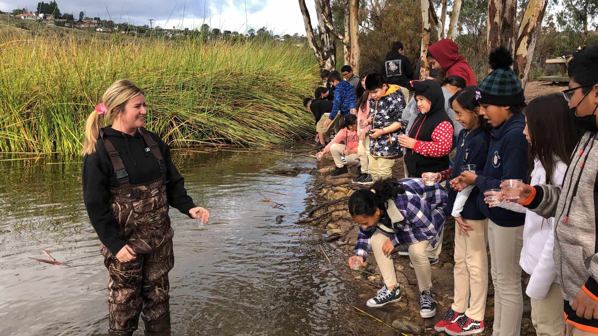 An educator stands in the River will students stand along the River bank preparing to release trout into the River.